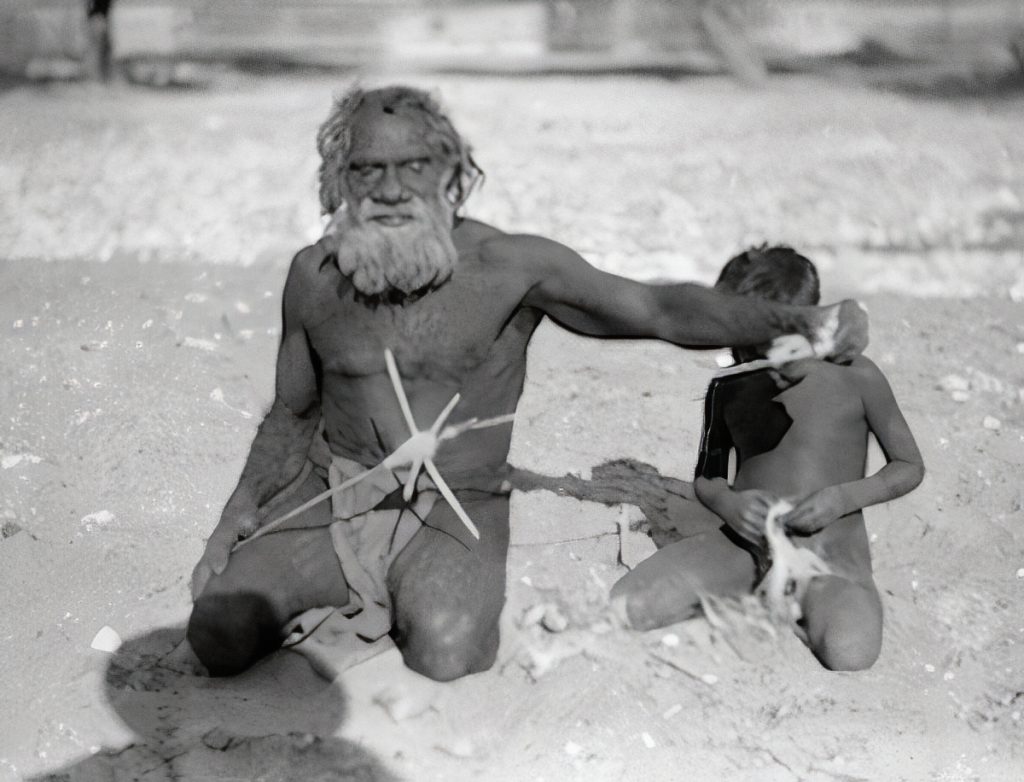 A Wangkangurru man spinning string with a wooden cross-piece spindle. The fibres were obtained by smashing up dry grass stalks. 1920s.